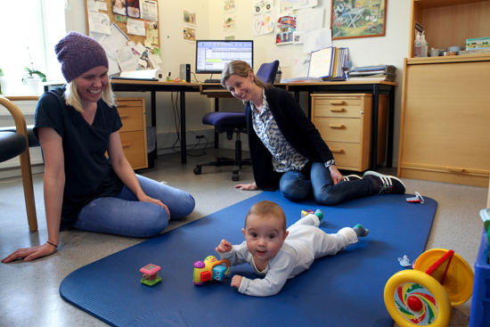 Kvinne og helsesykepleier observerer baby som ligger på magen på matte og leker 