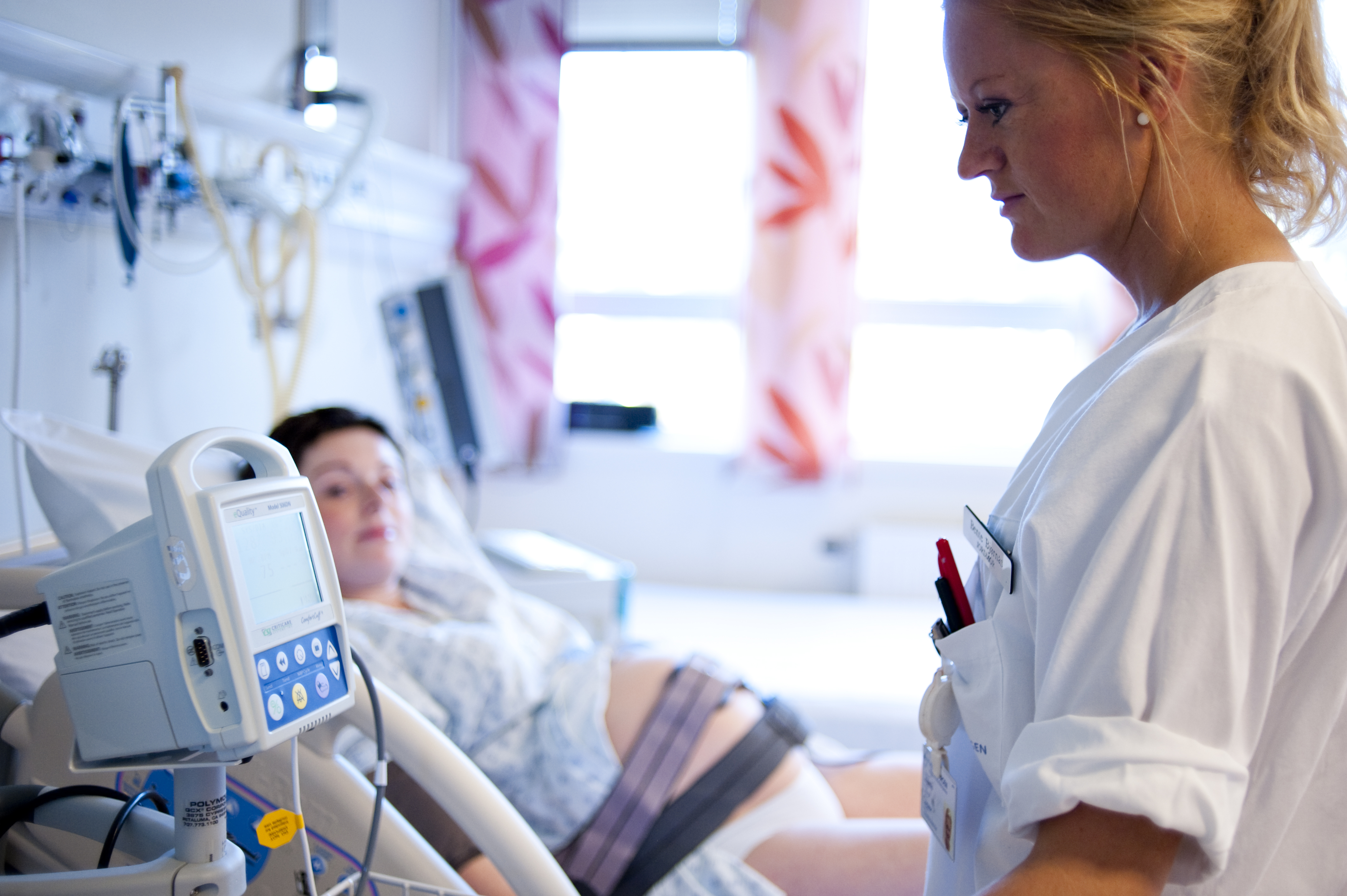 Gravid kvinne ligger i en sykehusseng med elastiske bånd med elektroder rundt magen, mens en sykepleier leser av resultatene på en CTG-maskin (cardiotokografi) som måler barnets hjerteslag.