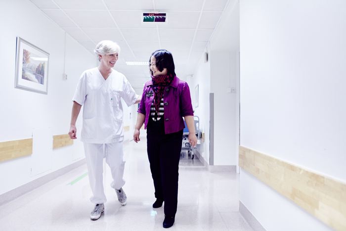 Smilende sykepleier følger pasient nedover sykehuskorridor.